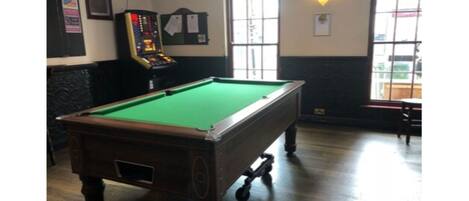 A pool table in a room with a wooden floor, a jukebox, and windows with cross-shaped decorations.