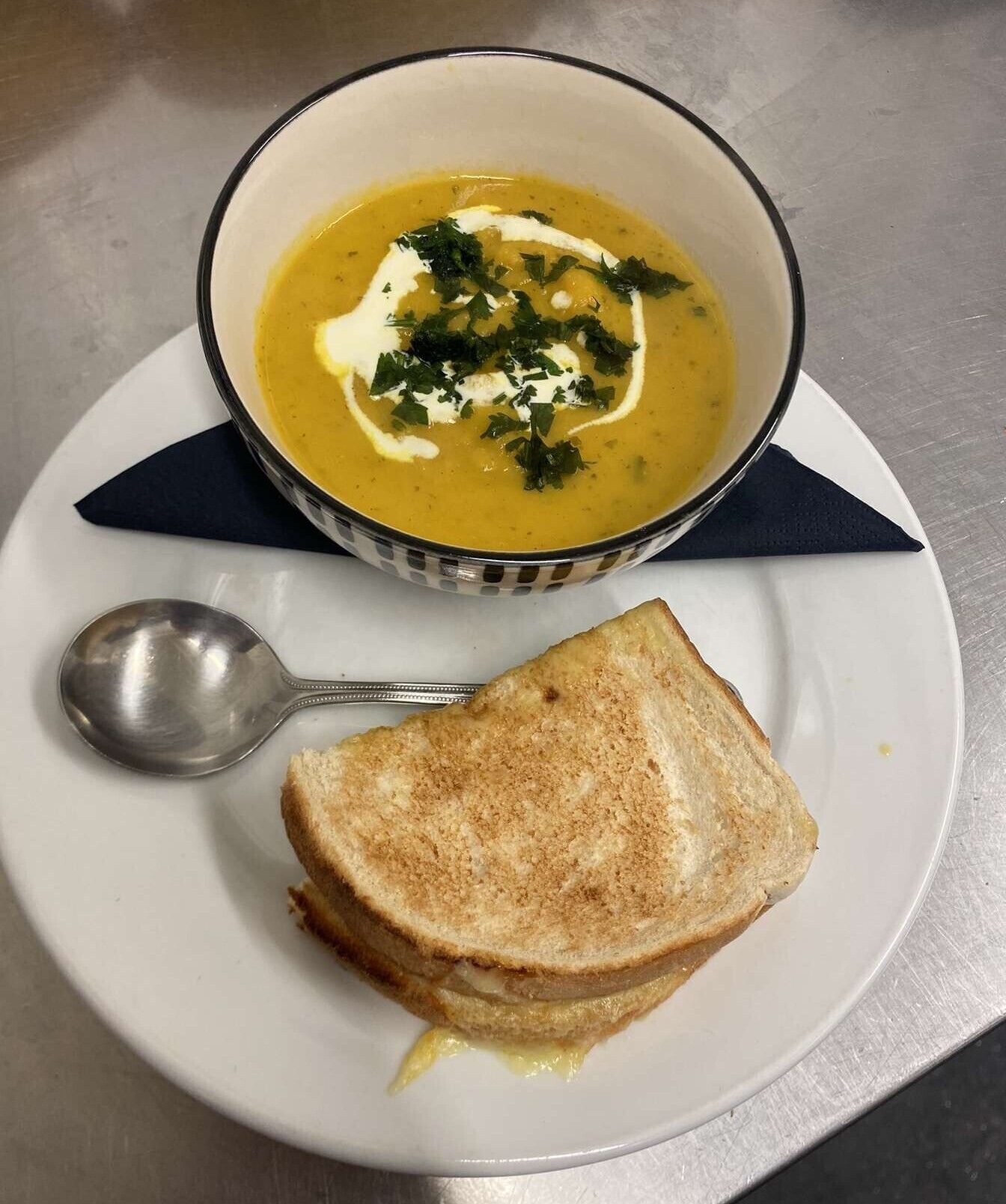 A bowl of soup with a side of grilled cheese sandwich and a spoon on a plate.