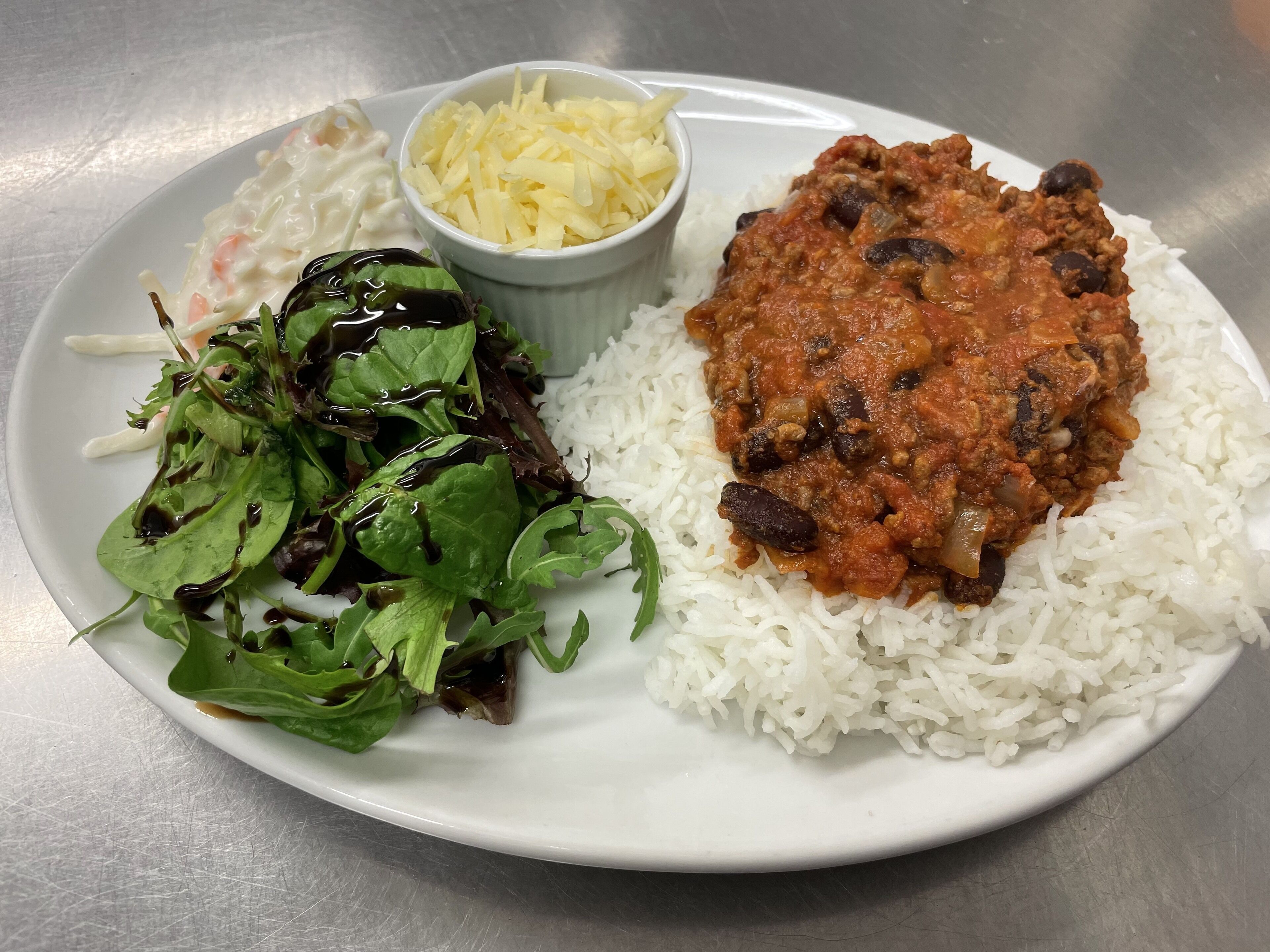 A plate with white rice, a meat-based dish, a side salad, and a small bowl of shredded cheese.