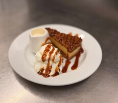 Un plato de postre con una porción de tarta, una bola de helado y un recipiente pequeño de salsa.