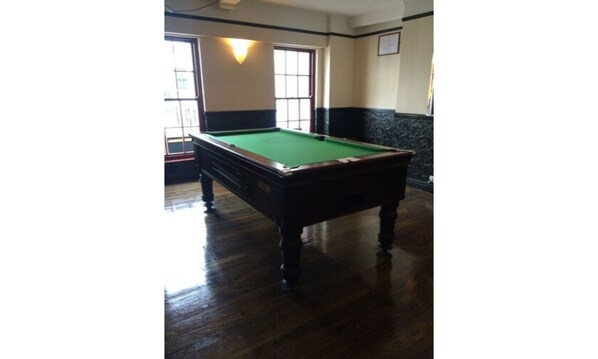 A pool table in a room with wooden flooring, large windows, and a dark wall with a light fixture.