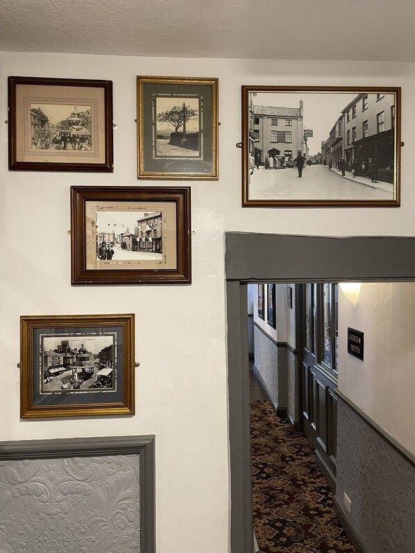 A hallway with framed black and white photographs on the wall, a sign indicating ladies and gentlemen, and a carpeted floor. - The Kings Arms (North Walsham)