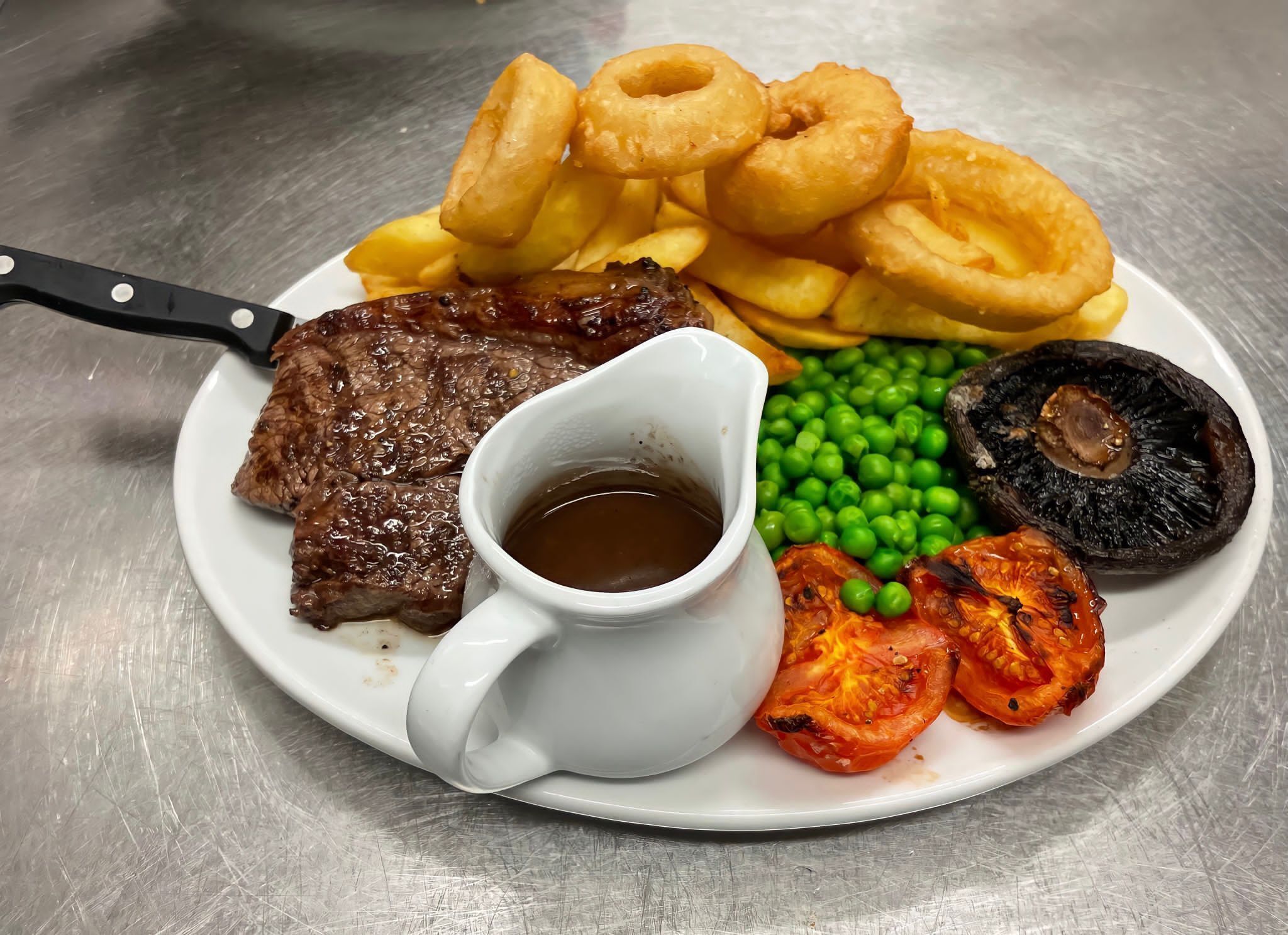 A plate with steak, onion rings, fries, peas, grilled tomatoes, and a gravy boat.
