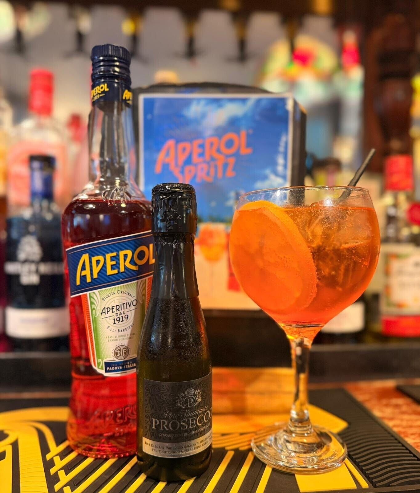 A bar setting with bottles of Aperol and Prosecco, a cocktail glass with an orange slice, and a blue sign in the background.