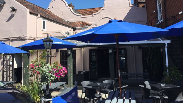 An outdoor seating area with blue umbrellas, wicker furniture, and a brick building in the background. - The Kings Arms (North Walsham)
