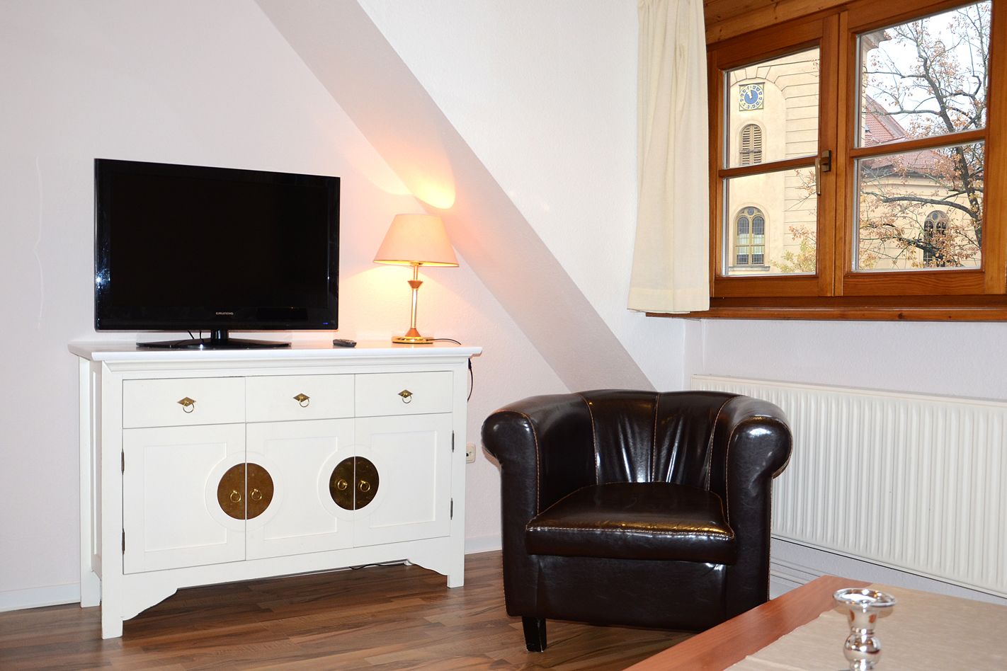 Una sala de estar con un televisor sobre un mueble blanco, un sillón de cuero y una ventana con vistas a un edificio.