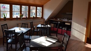 A dining area with wooden tables and chairs, a kitchen in the background, and a view through large windows. - FRITZ Aparthotel Potsdam (Potsdam)