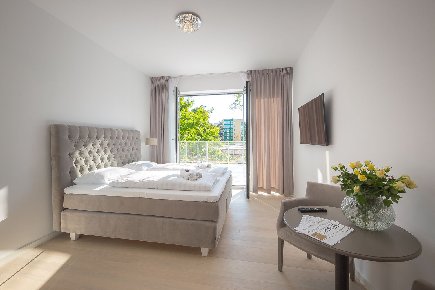 Un dormitorio moderno con una cama grande, un balcón con cortinas, una mesita con un jarrón y un televisor en la pared.