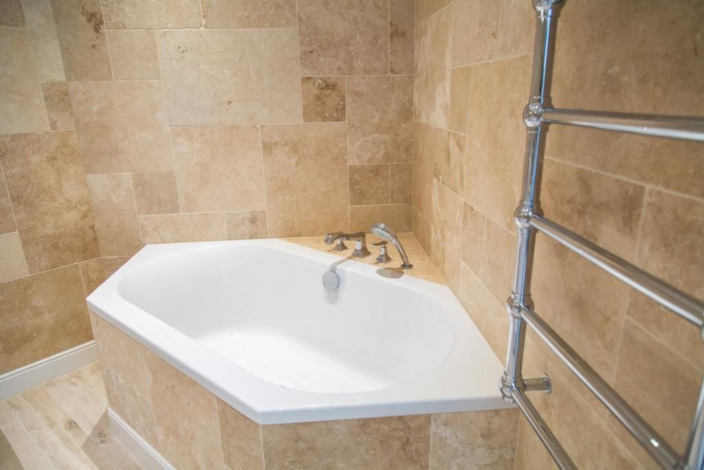 Un baño en esquina con una bañera blanca, azulejos color beige y un perchero de metal.