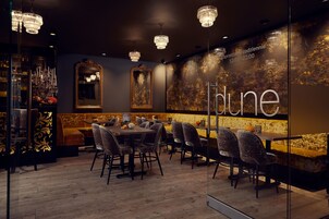 A restaurant interior with wooden flooring, multiple tables and chairs, chandeliers, and a wall with the word 'dune' and a clock indicating operating hours.
