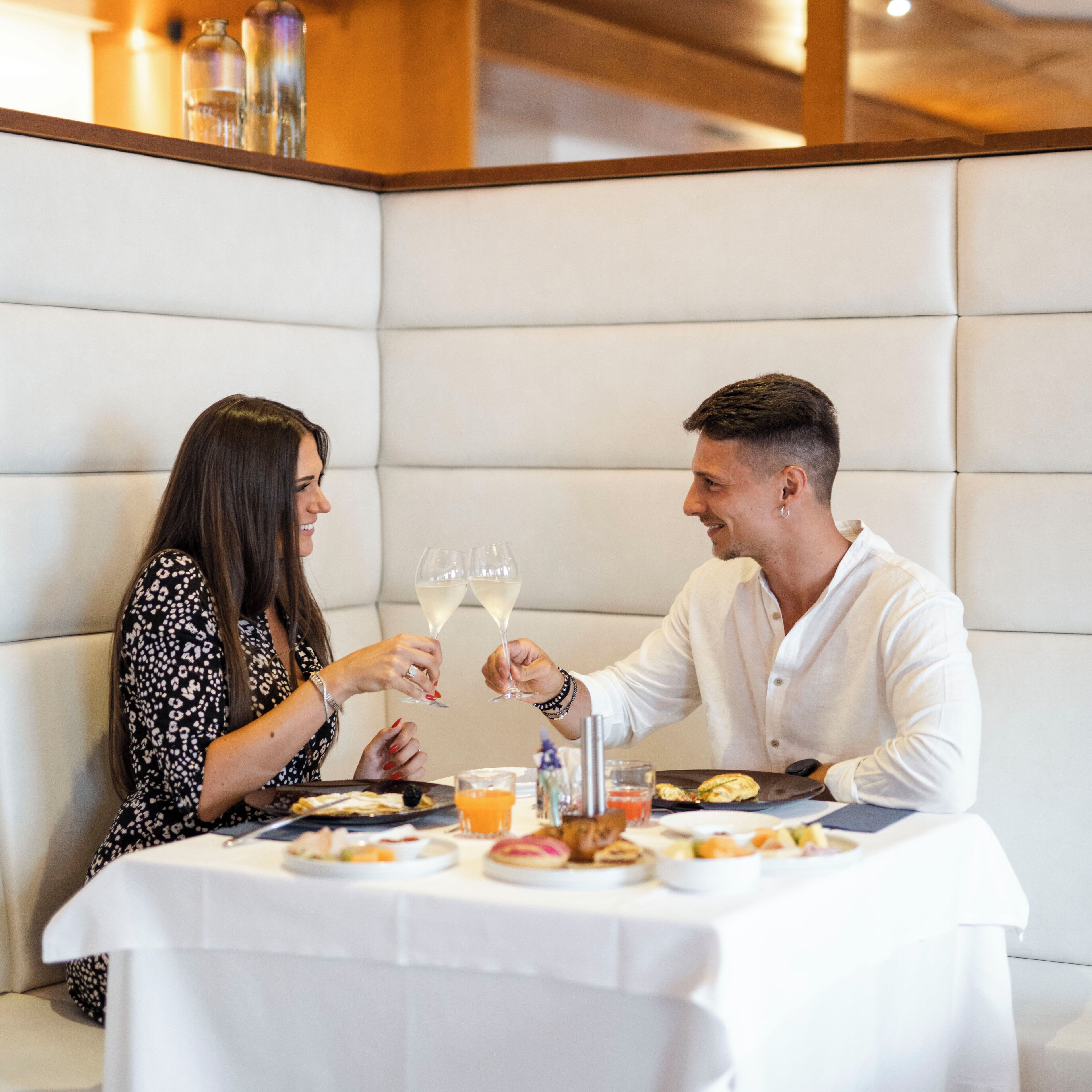 Una pareja sentada a una mesa con mantel blanco, sosteniendo copas de vino. Hay platos de comida y un vaso de jugo de naranja sobre la mesa.