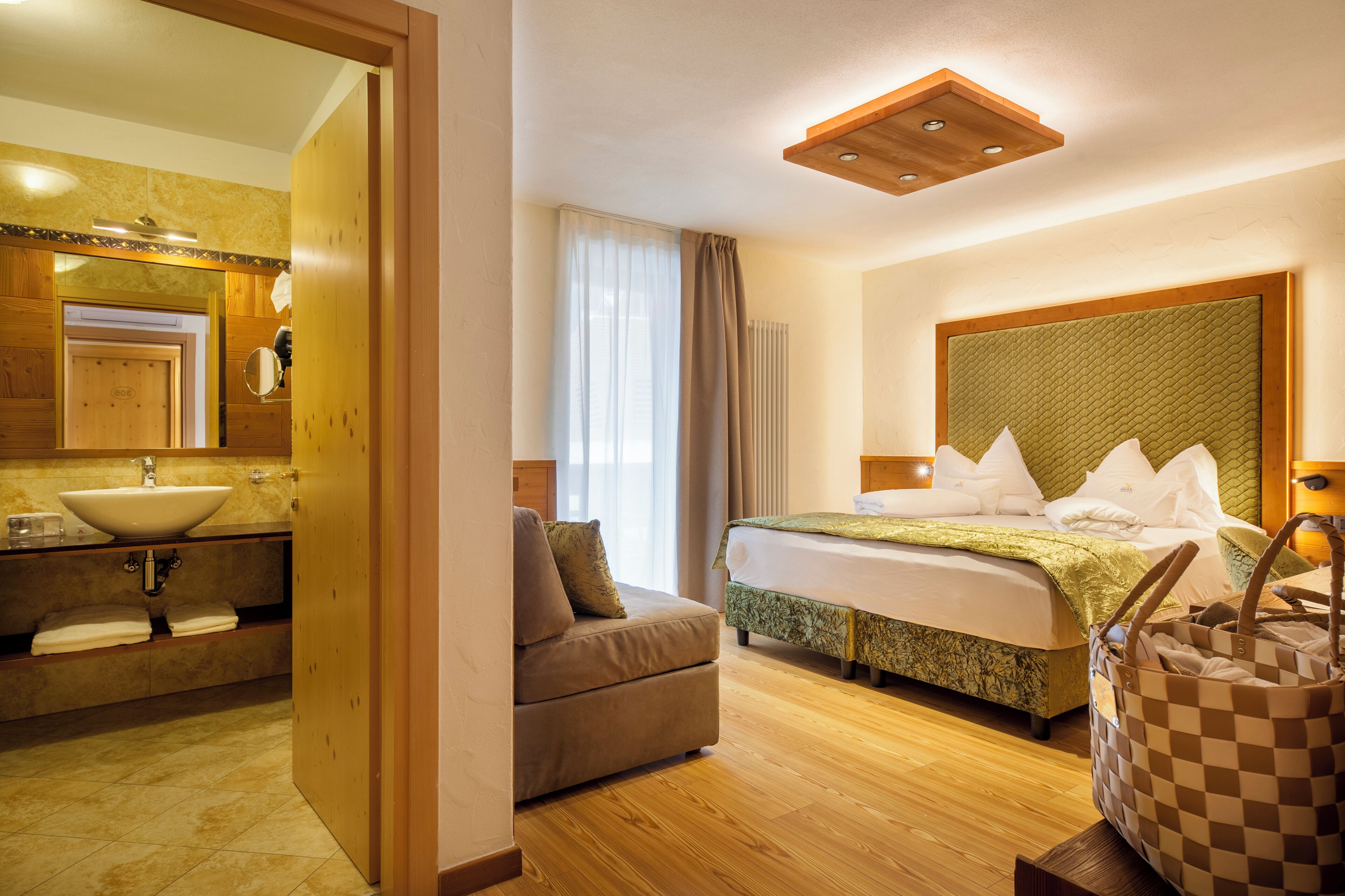 Ein Hotelzimmer mit Holzboden, einem Bett mit grünem Kopfteil, einer Couch, einem Badezimmer mit Waschbecken und Spiegel sowie einer karierten Tasche auf einem Tisch.