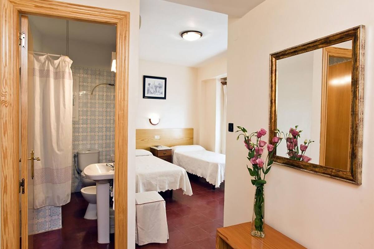 A hotel room with a wooden door, a shower, a toilet, a sink, two beds with white linens, a bedside table, a mirror, and a vase with flowers.