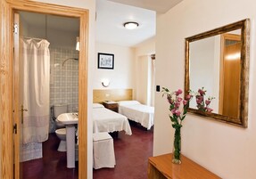 Habitación de hotel con puerta de madera, ducha, inodoro, lavamanos, dos camas con ropa blanca, una mesita de noche, un espejo y un florero con flores.
