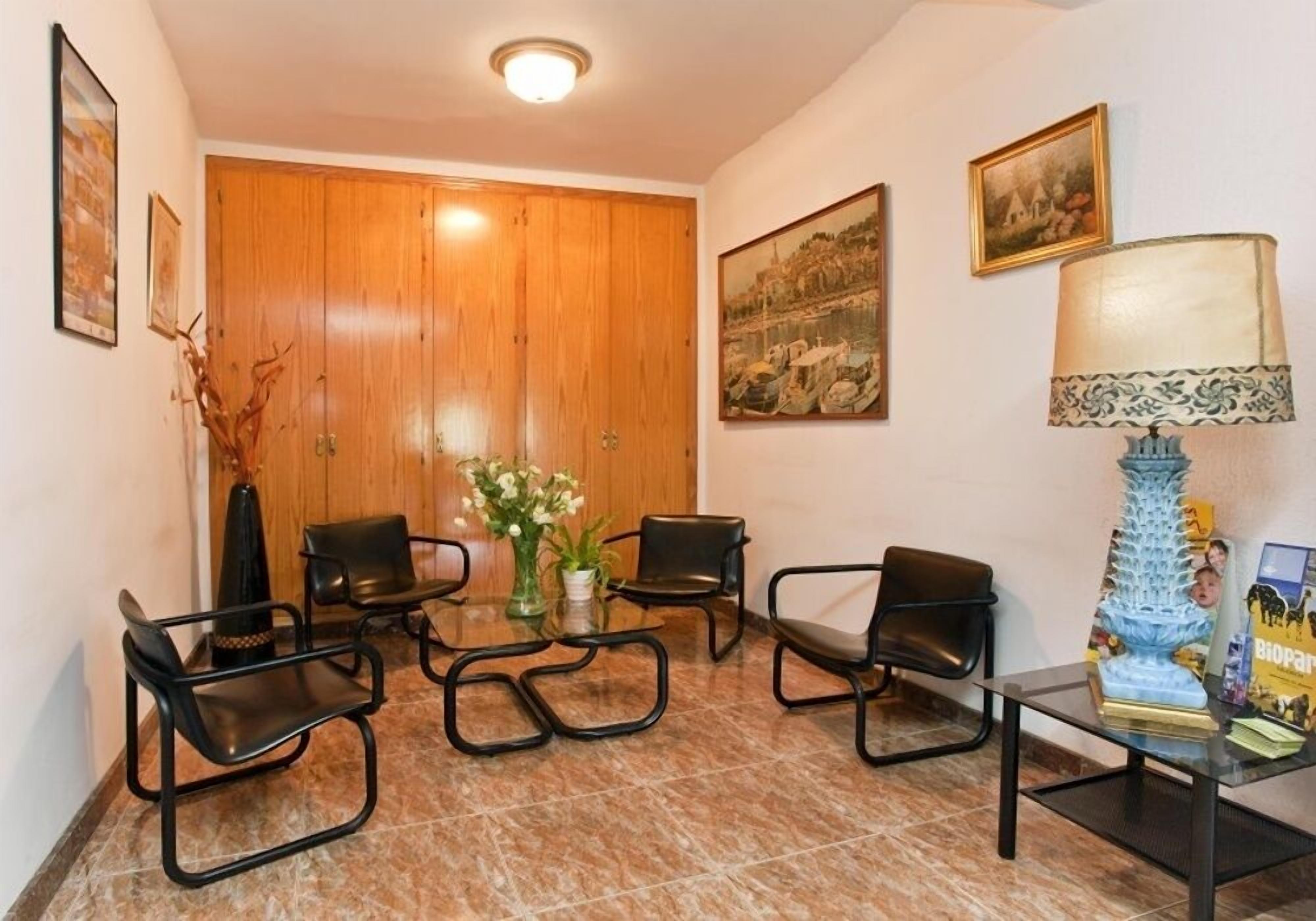 A lobby with a wooden door, black chairs, a glass table, a vase with flowers, and a lamp.