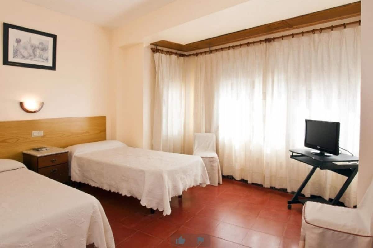 A hotel room with two beds, a desk with a computer, a chair, a window with curtains, and a framed picture on the wall.