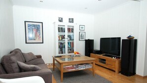 A living room with a brown sofa, a glass-top coffee table, a bookshelf, a TV, and a black speaker. - Solferie Holiday Home - Korsvikkilen (Kristiansand)