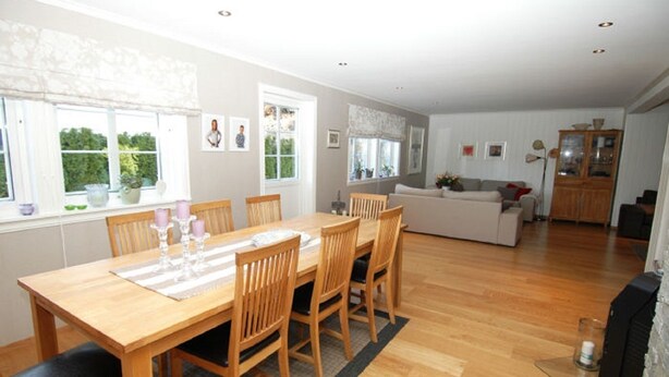 A spacious living area with a wooden dining table, chairs, and a sofa. There are windows with curtains, a fireplace, and a kitchen area in the background. - Solferie Holiday Home - Korsvikkilen (Kristiansand)