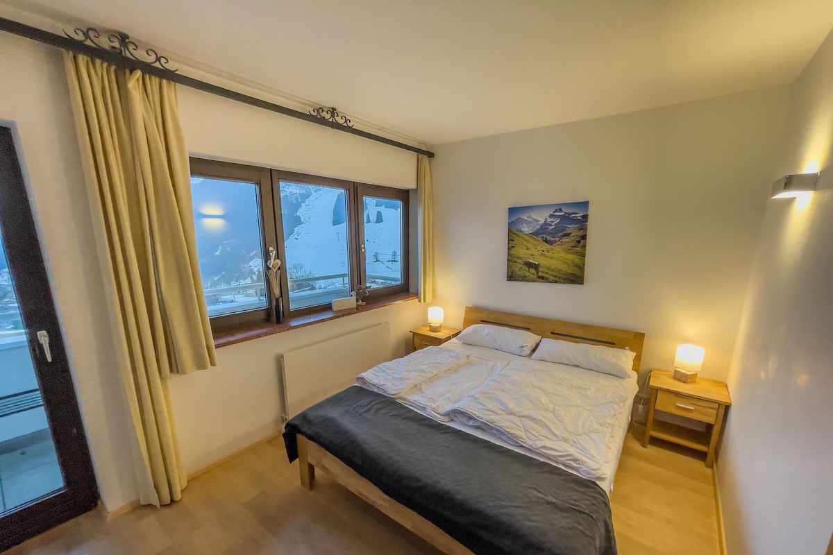 A bedroom with a bed, two bedside tables with lamps, a window with curtains, a radiator, and a painting on the wall.