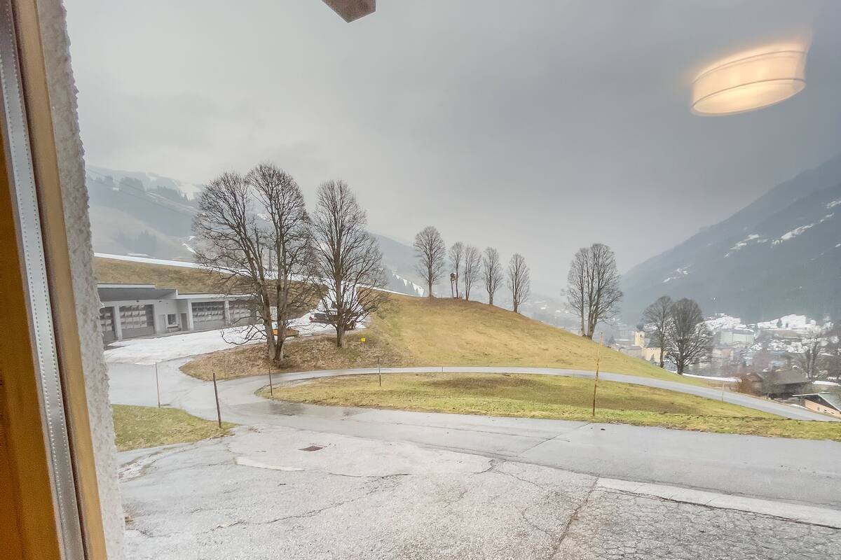 A view through a window of a parking area with a grassy hill and bare trees, leading to a building with a flat roof.