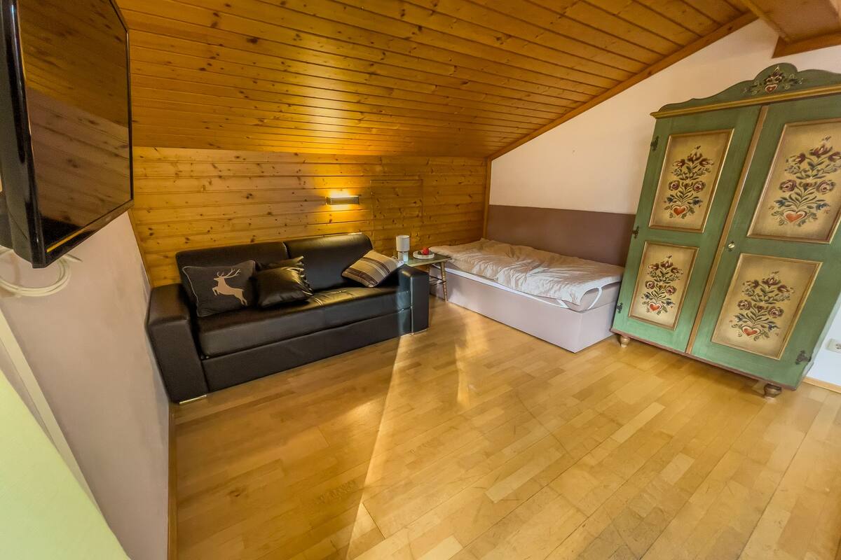 A cozy attic bedroom with a wooden ceiling, a small sofa, a single bed, and a green cabinet with floral designs.