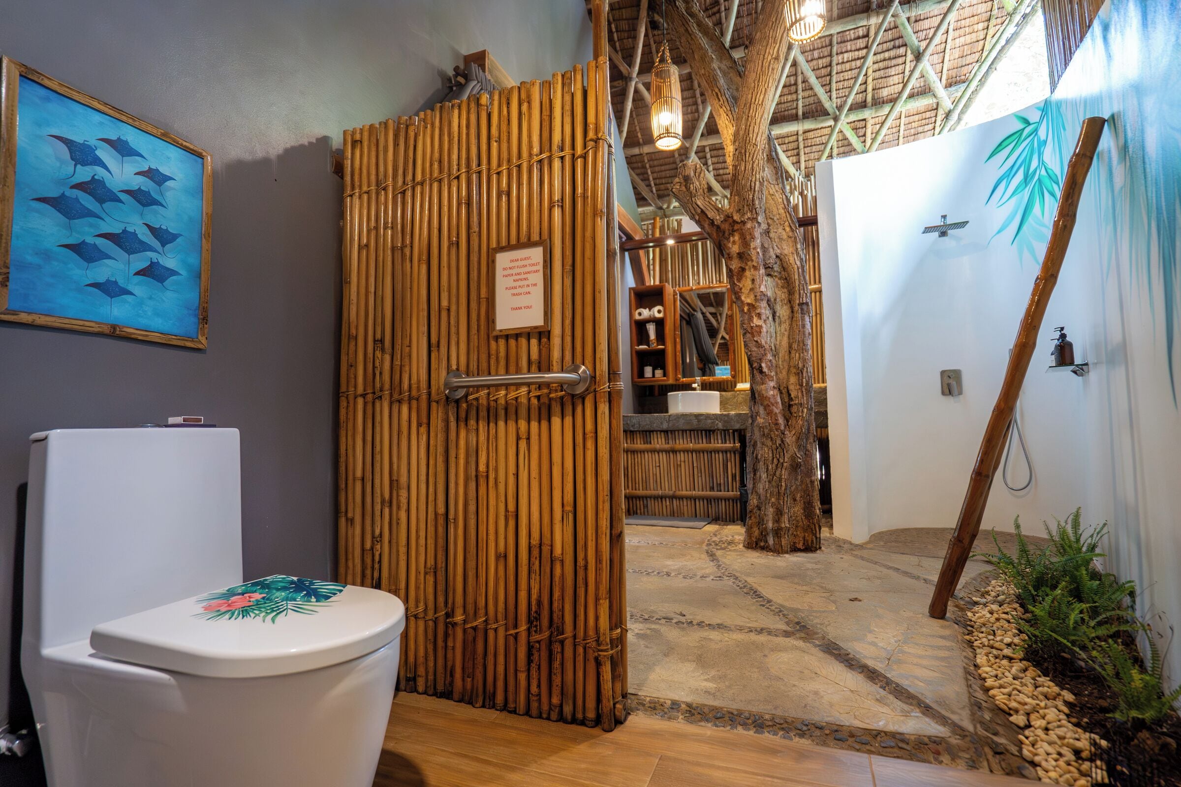 Un baño con puerta de bambú, inodoro con asiento floral, un cuadro en la pared y zona de ducha con una planta de bambú.
