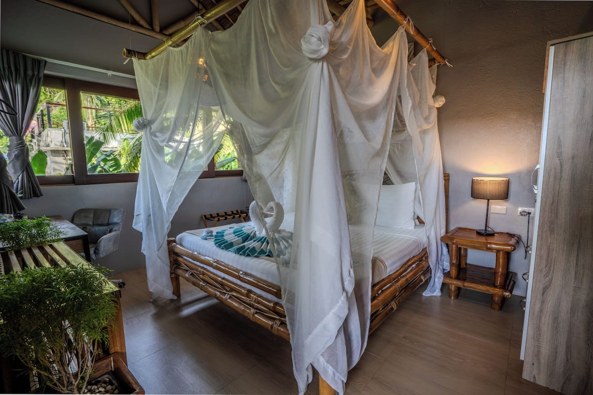 A room with a bed under a canopy, a bedside table, a lamp, and a view of greenery outside.
