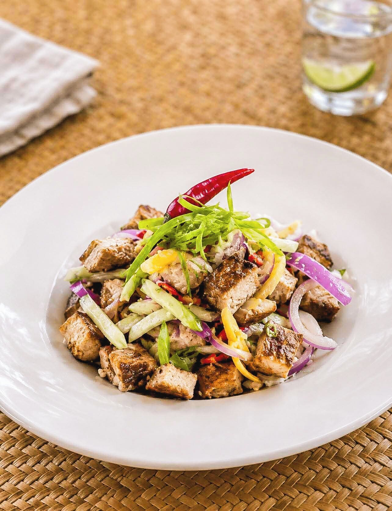 A white plate with a dish of grilled meat and vegetables, garnished with green herbs and a red chili.