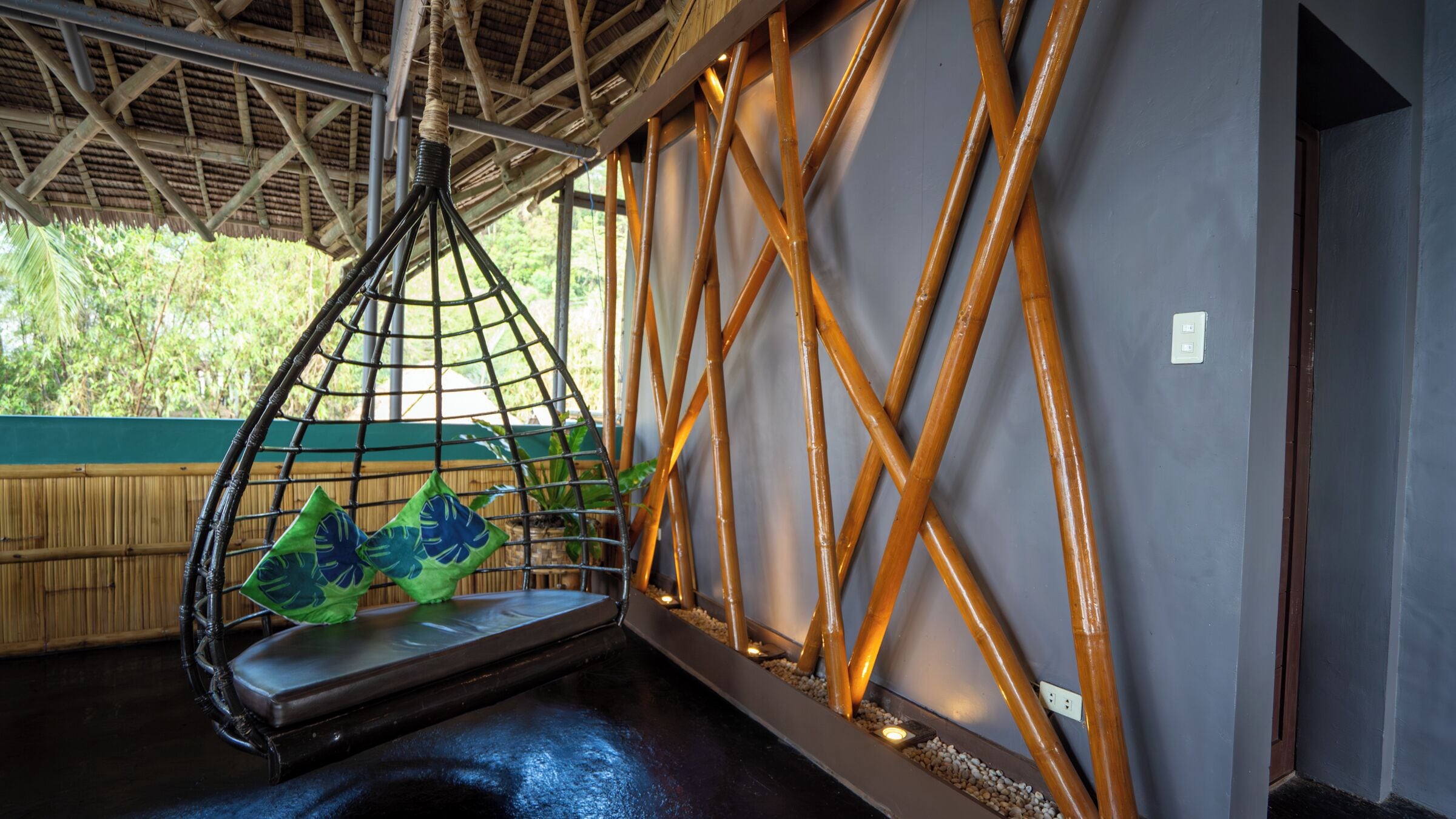 Quarto com cadeira suspensa, parede de bambu e vista para o verde.
