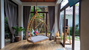 A room with a hanging rattan chair, a wooden floor, and a view of a garden through large windows.