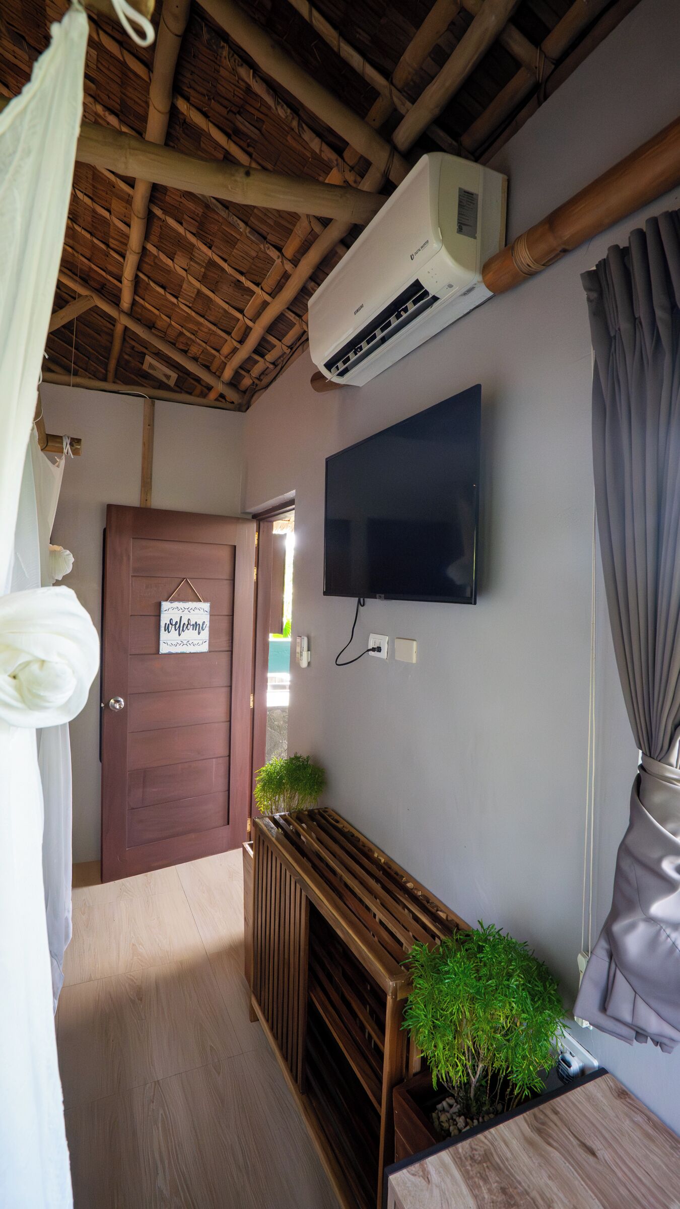 Una habitación con techo de madera, televisor montado, puerta con letrero de 'welcome', banco con plantas y ventana con cortinas.