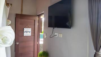 Una habitación con techo de madera, televisor montado, puerta con letrero de 'welcome', banco con plantas y ventana con cortinas.