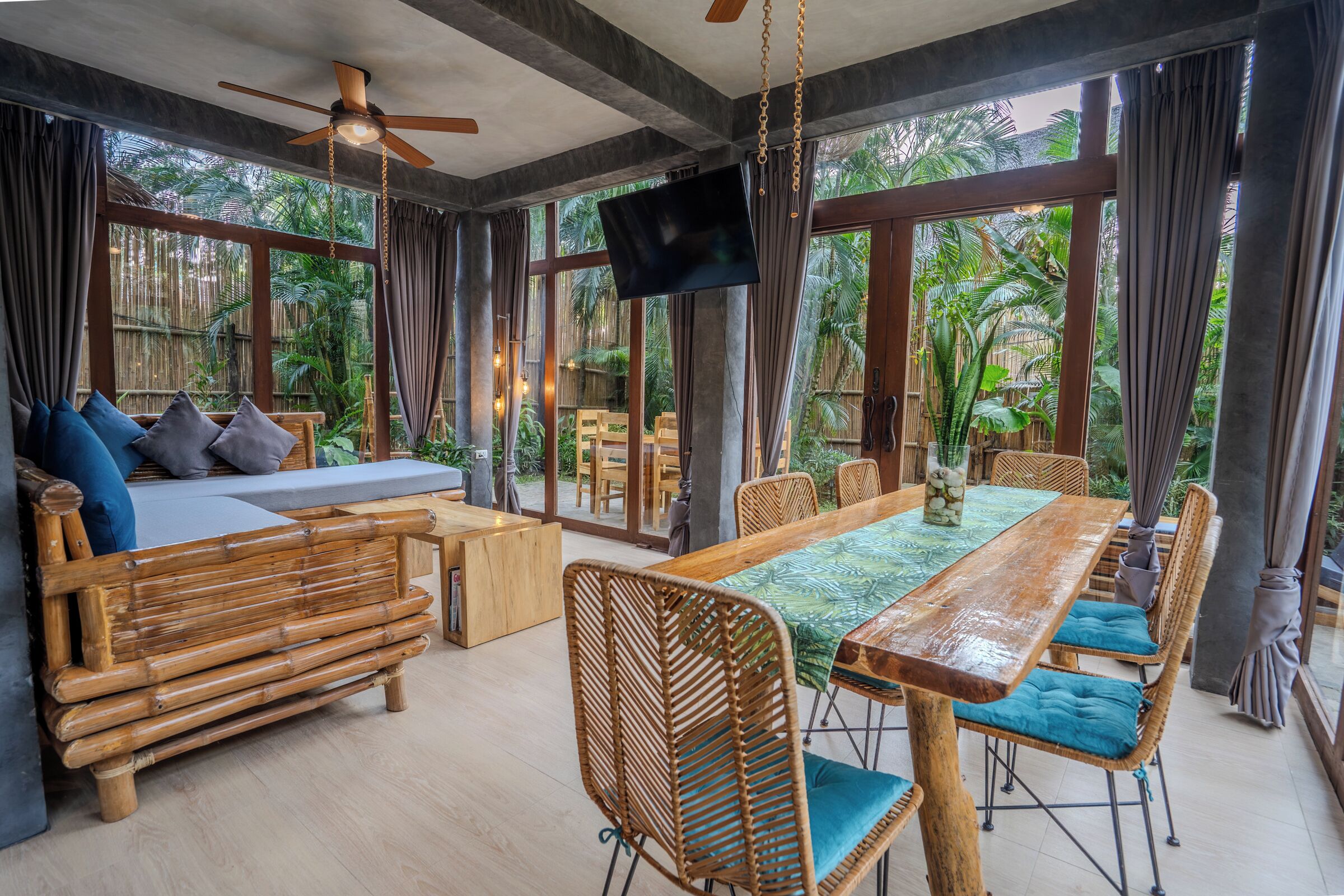 Una habitación con un ventanal grande, ventilador de techo, televisor, mesa de comedor de madera y sofá con cojines.