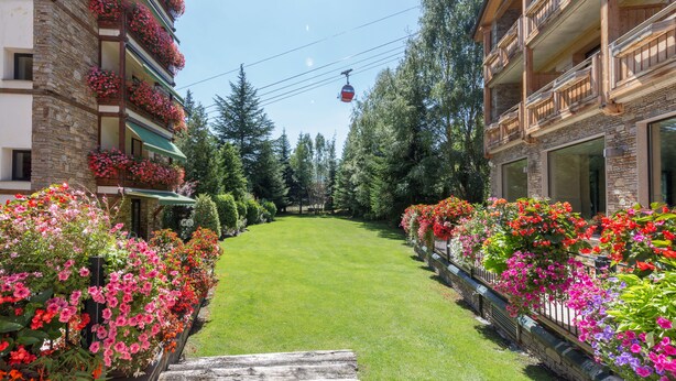 A hotel corridor with a green lawn, flower boxes, and a cable car in the background.