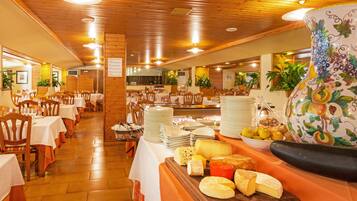 A restaurant with wooden interiors, multiple tables, and a buffet with cheese and fruits.