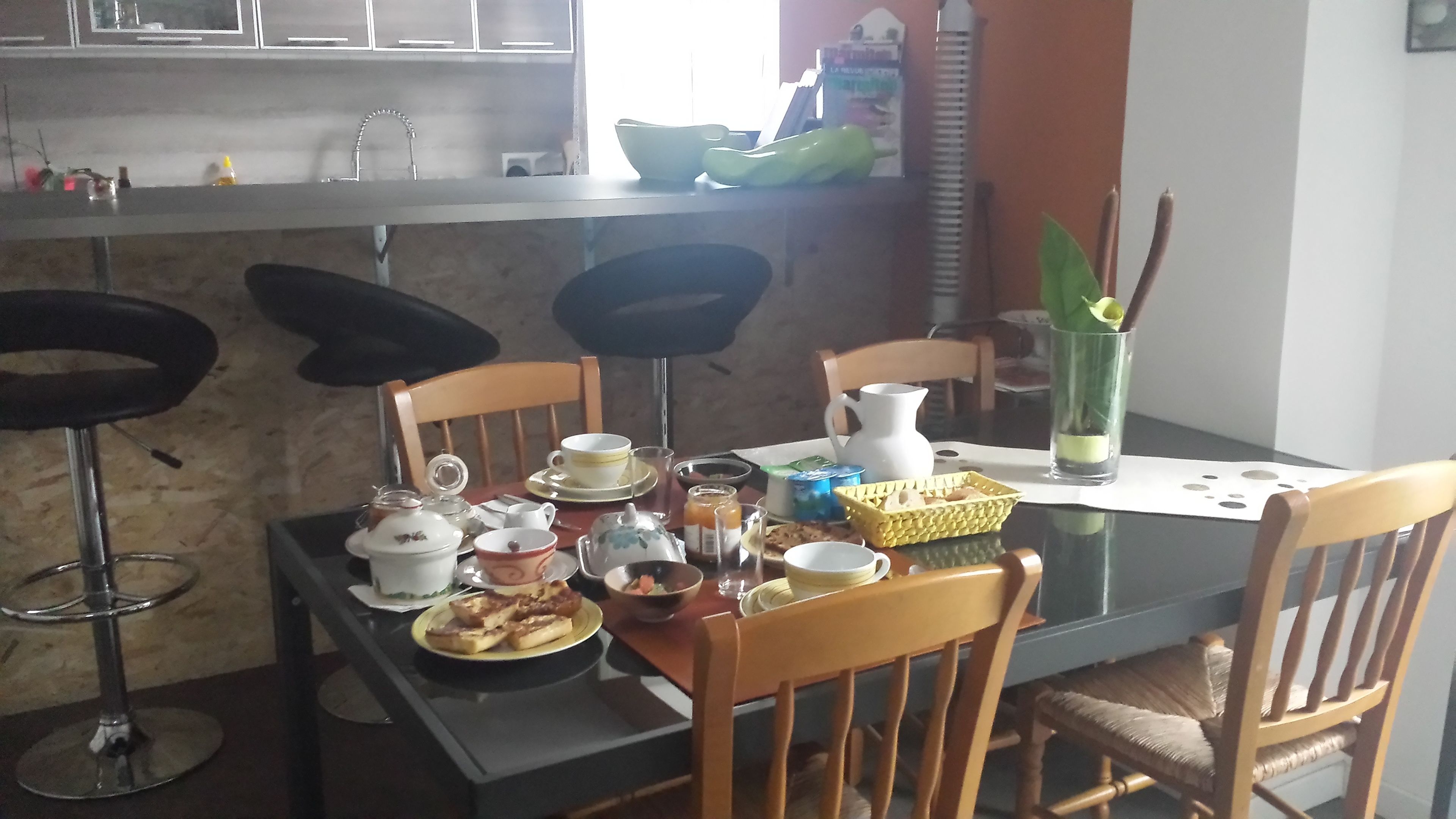 Une table à manger est dressée avec différents plats, tasses et un pichet. Des chaises sont disposées autour de la table, et on aperçoit un coin cuisine à l’arrière-plan.