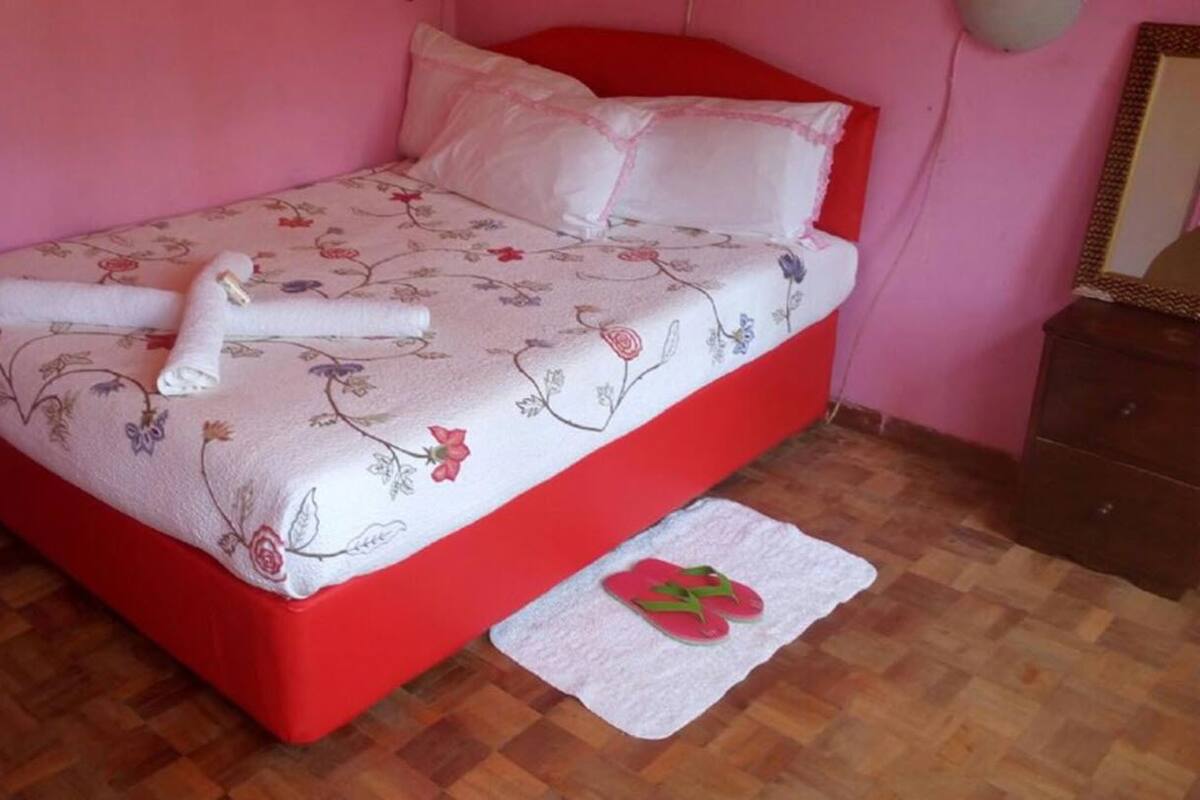 A bed with floral bedding and red headboard, a white towel, and a pair of flip-flops on a white mat.