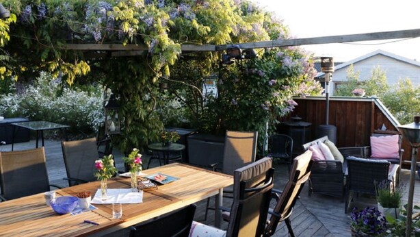 An outdoor patio area with wooden tables and chairs, surrounded by lush greenery and flowering plants. - Solferie Holiday Home Vardåsveien (Kristiansand)