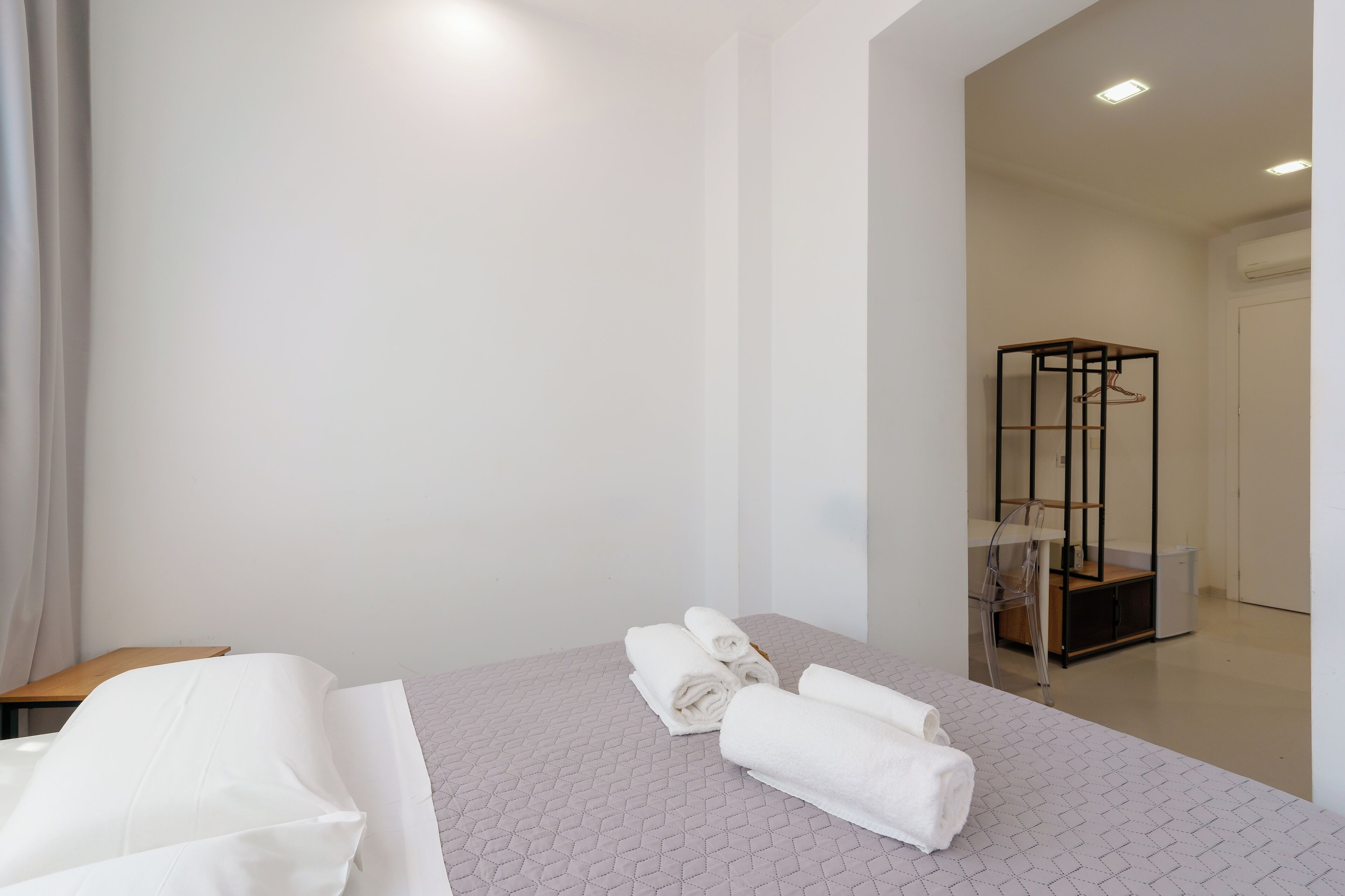 A hotel room with a neatly made bed, a small wooden table, and a metal-framed wardrobe.