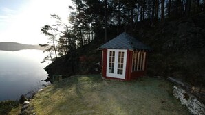 A small red cabin with a white door, surrounded by a grassy area and a wooden fence, near a body of water and forested hills.