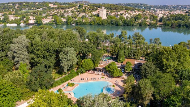 An aerial view of a resort with a large swimming pool surrounded by lush greenery, a tower, and a lake in the background. - Camping Du Pont D'Avignon (Avignon)