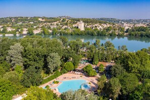 An aerial view of a resort with a large swimming pool surrounded by lush greenery, a tower, and a lake in the background. - Camping Du Pont D'Avignon (Avignon)
