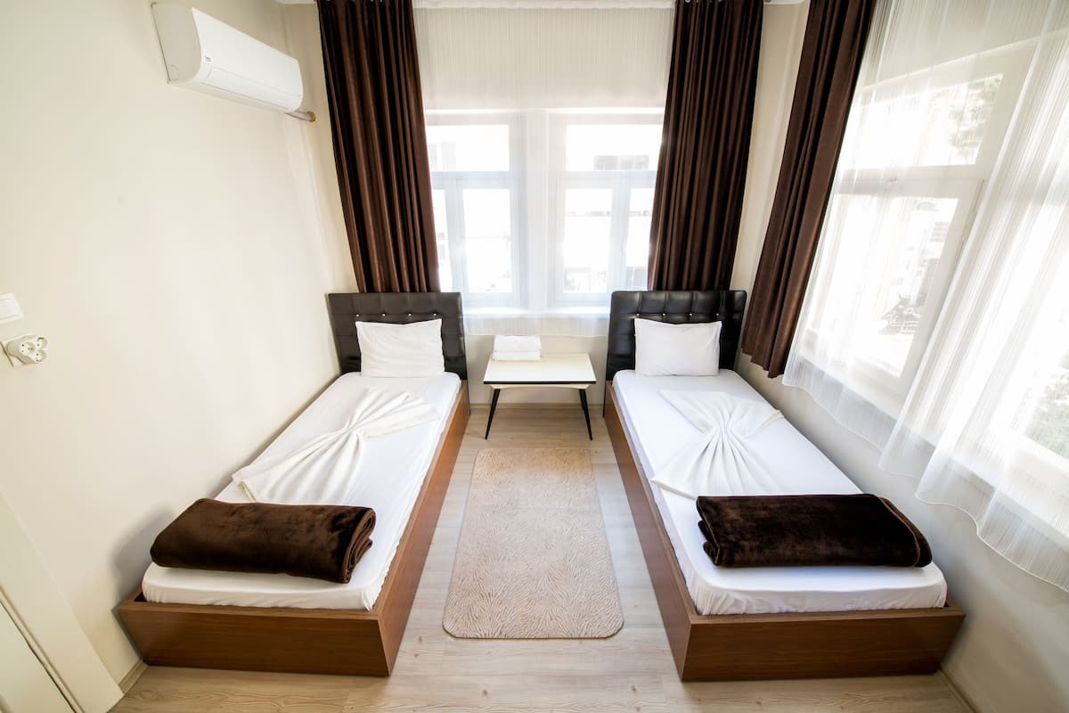 A room with two single beds, a small table, and a view through a window with curtains.