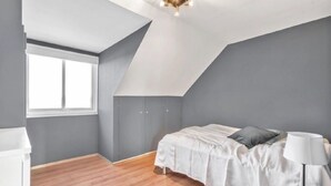 Ein Schlafzimmer mit schrägen Wänden, einem Fenster, einem Bett mit Kissen, einer Nachttischlampe und einem kleinen Tisch.