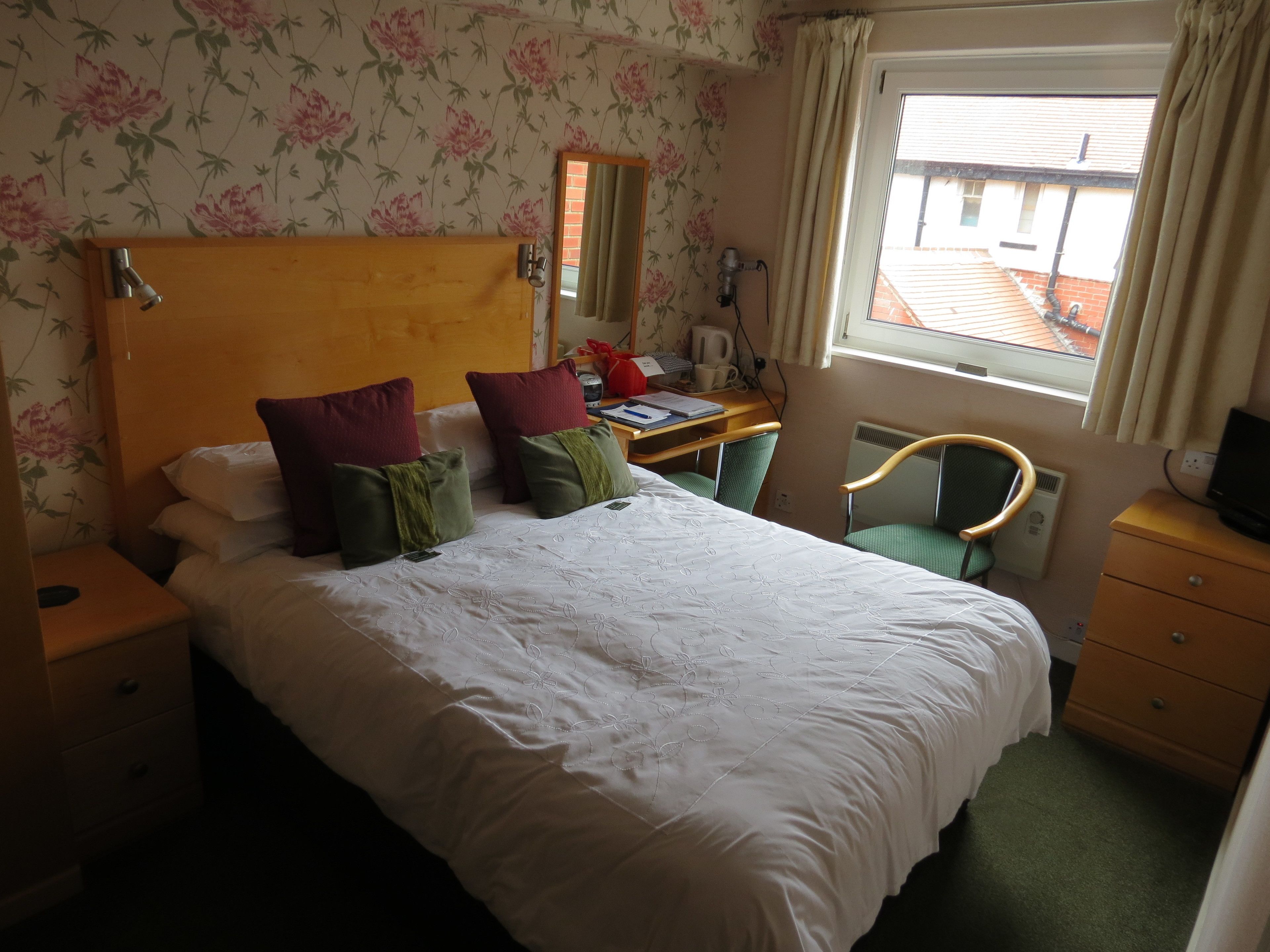 A hotel room with a large bed, a desk with a chair, a mirror, and a window with curtains.