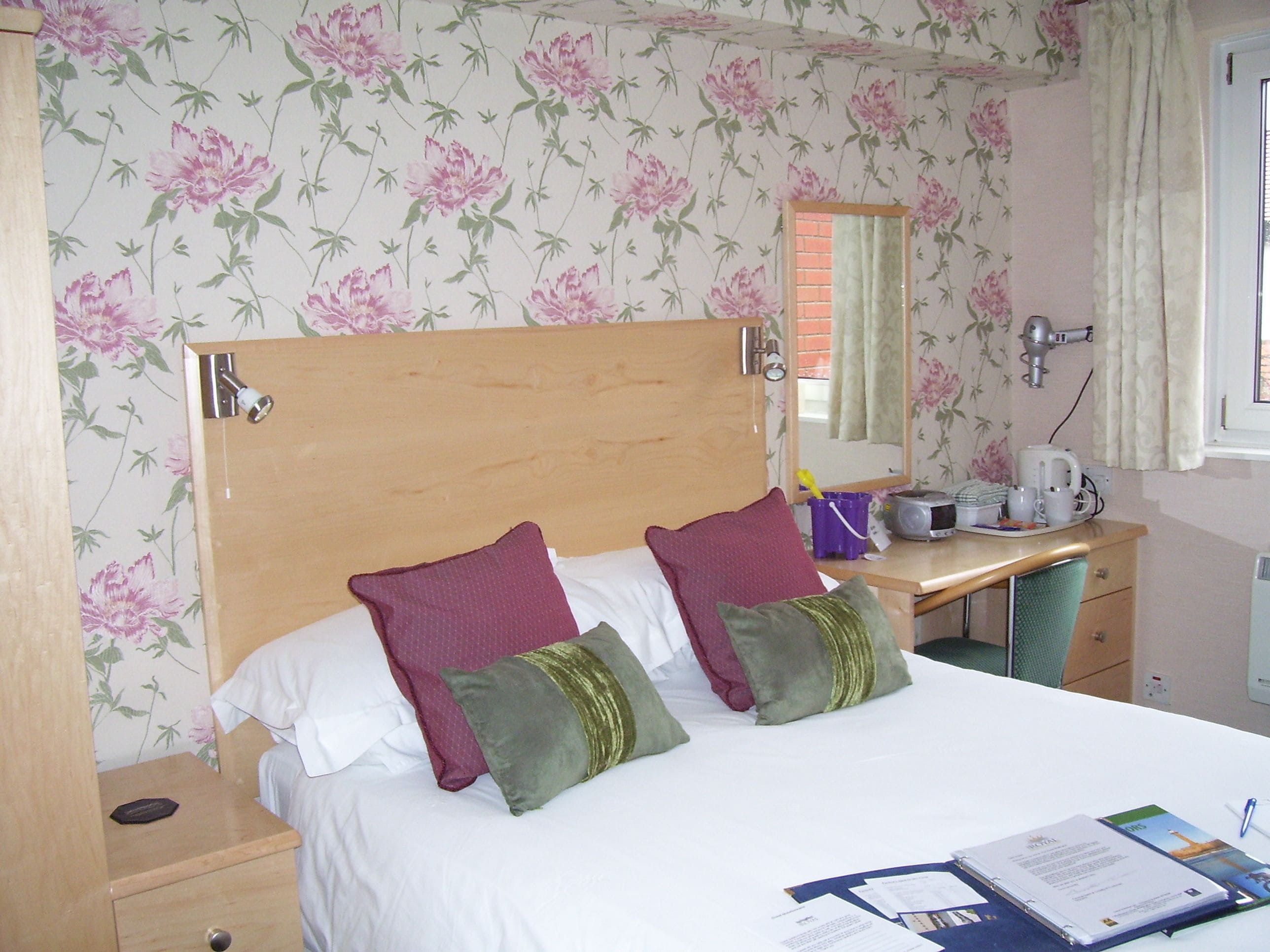 A hotel room with a wooden headboard, two pillows, a desk with a mirror, a kettle, and a window with curtains.