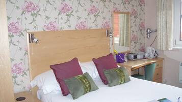 A hotel room with a wooden headboard, two pillows, a desk with a mirror, a kettle, and a window with curtains.