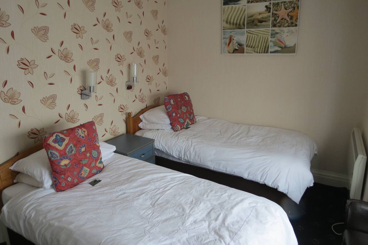 A room with two single beds, a small table, a wall-mounted lamp, and a decorative wall panel.