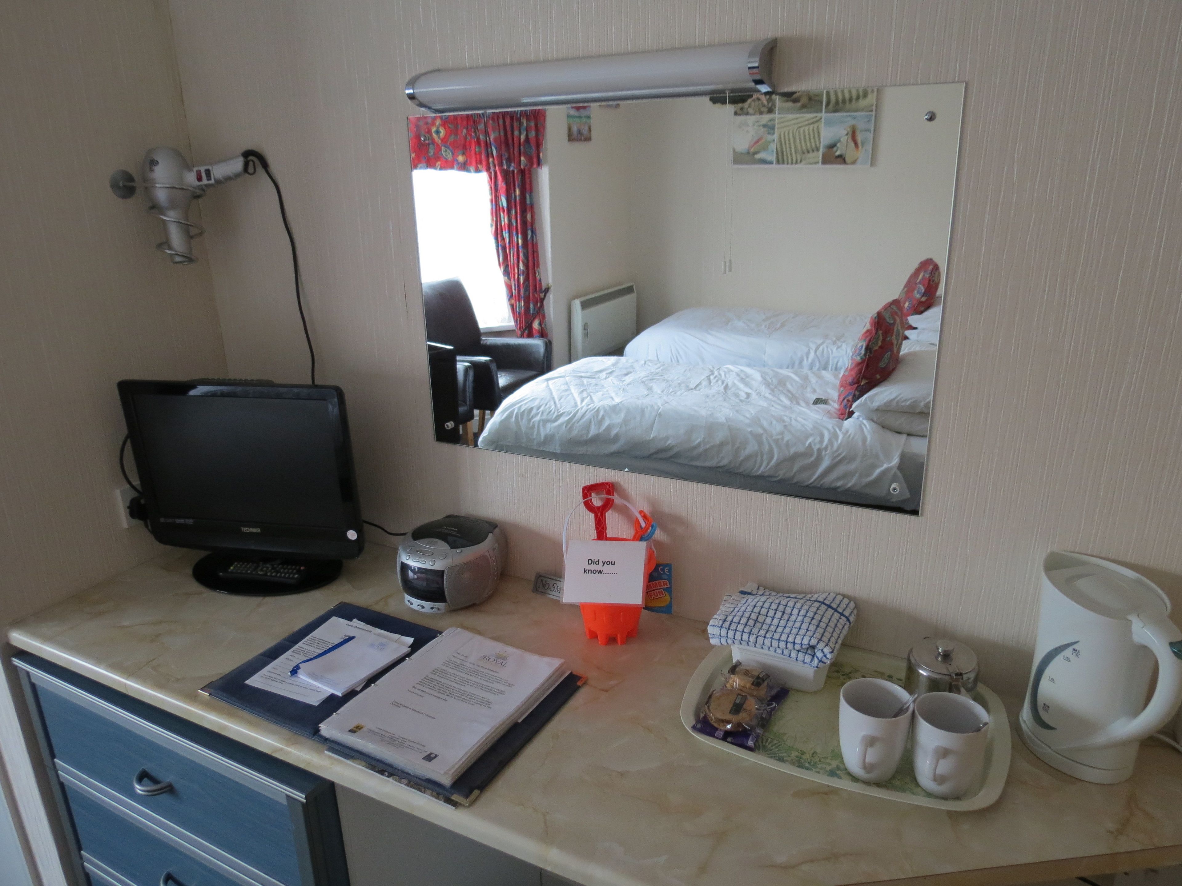 A hotel room with a bed, a desk with a computer, a mirror, a kettle, and a tray with cups and snacks.
