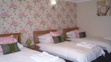 A hotel room with three single beds, each with white linens and green and pink pillows. There is a green lamp on the nightstand beside each bed.